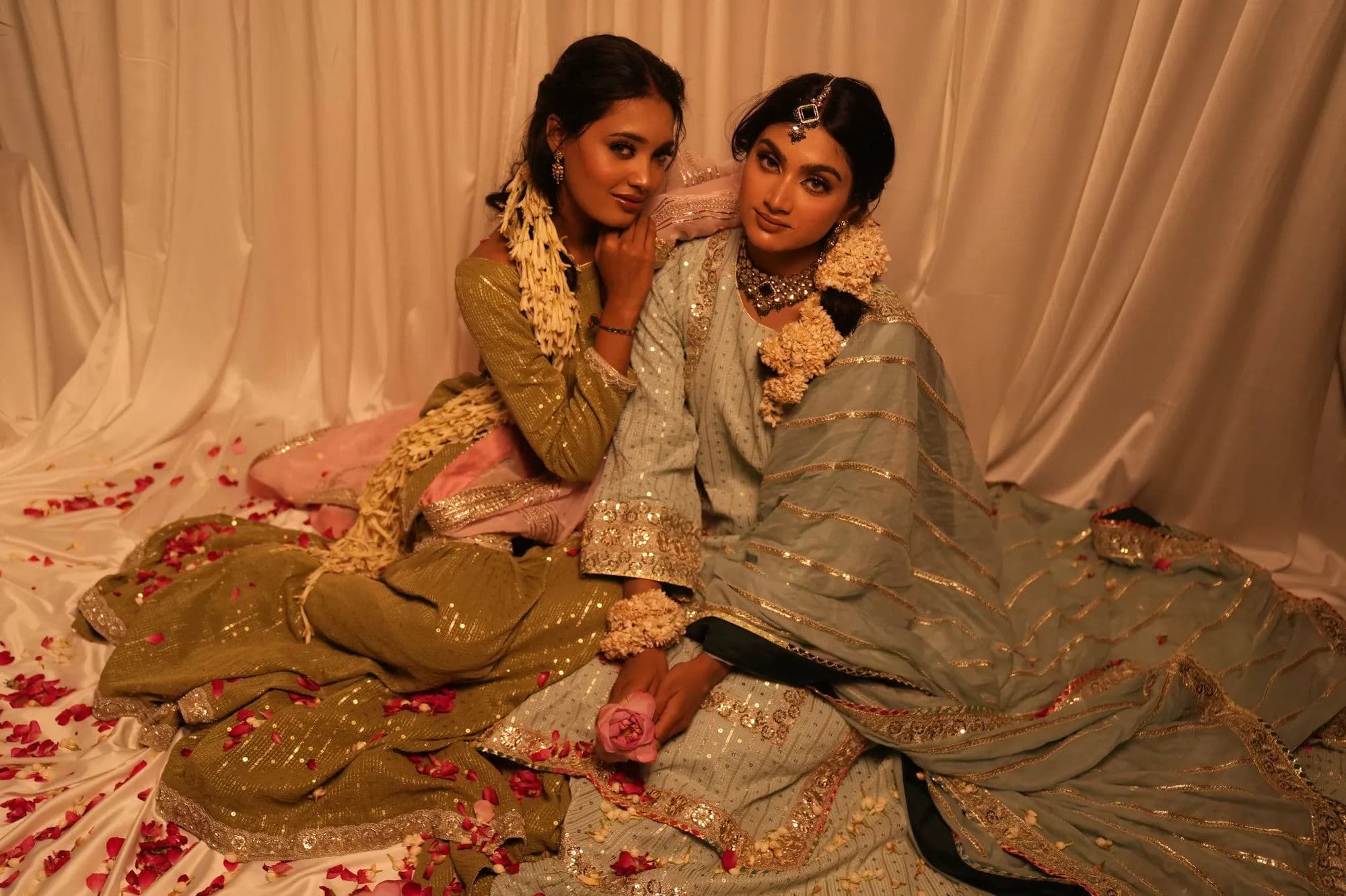 Two women in festive Sheshri couture, an olive sharara and a sage lehenga both worked in gold, seated together among scattered rose petals.
