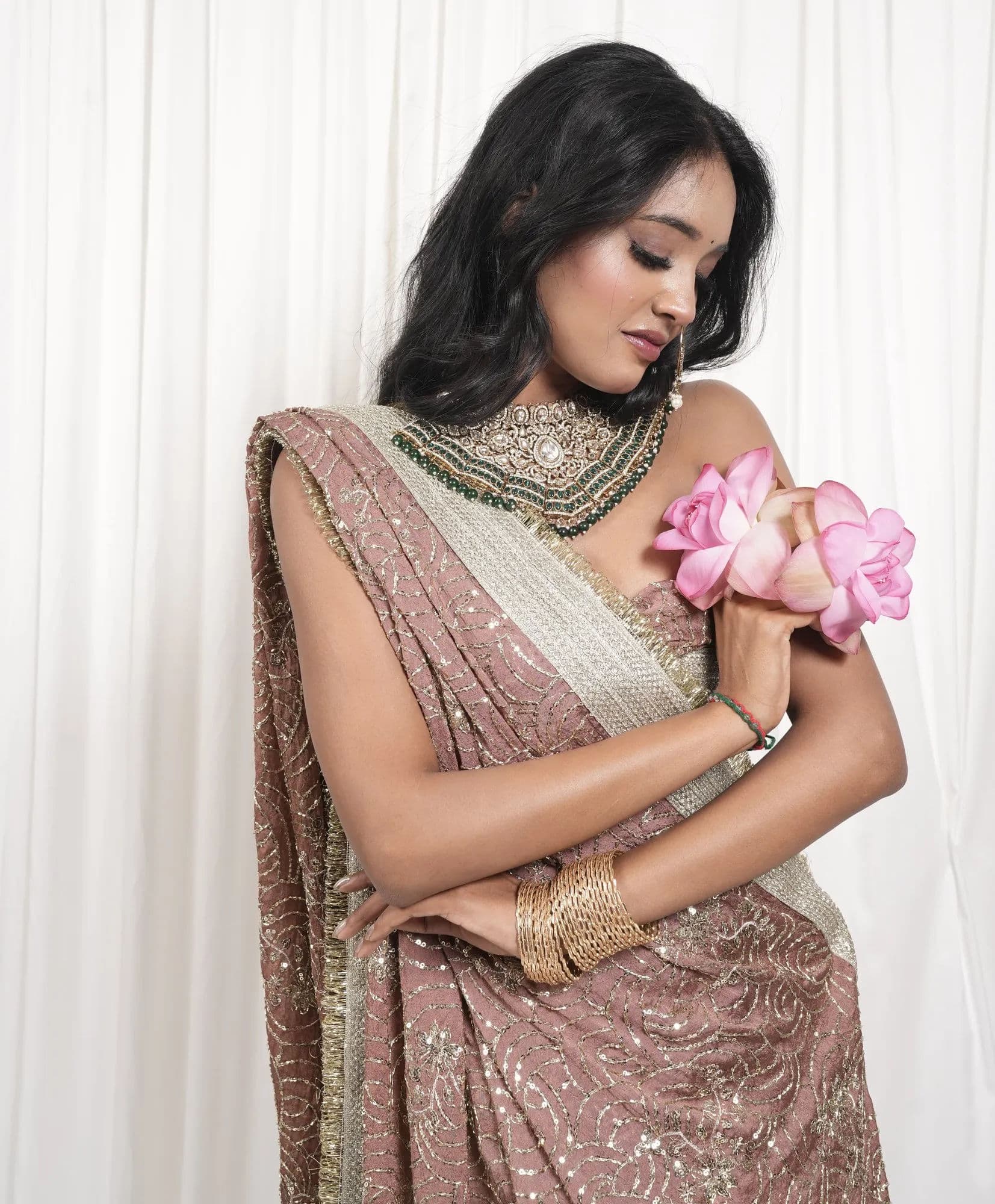 A blush and gold embroidered saree, worn with a beaded neckpiece and a cluster of pink blooms.